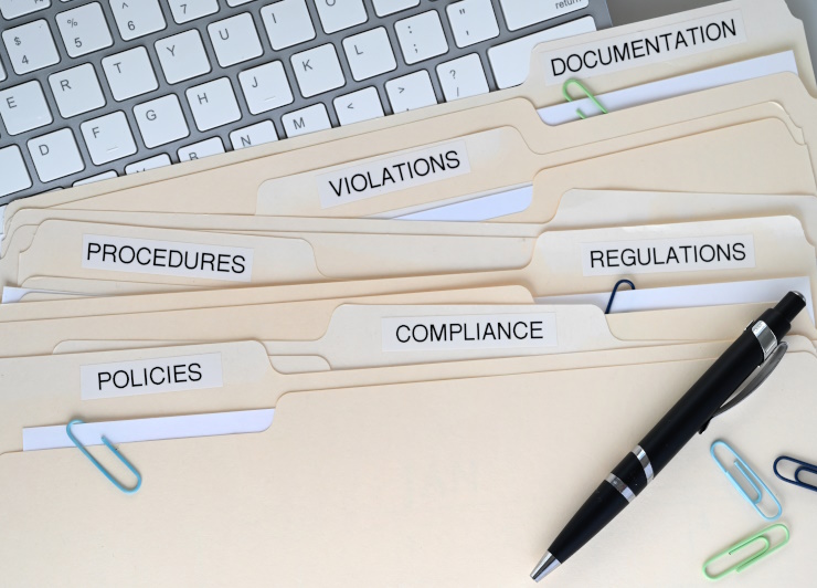 Folder of documents in front of a keyboard with words like polices, compliance, regulations on the header tabls.